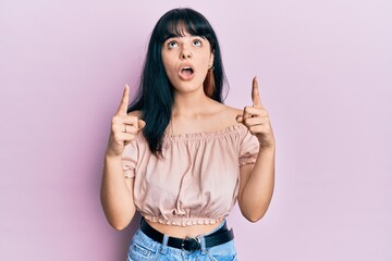 Young hispanic girl wearing casual clothes amazed and surprised looking up and pointing with fingers and raised arms.