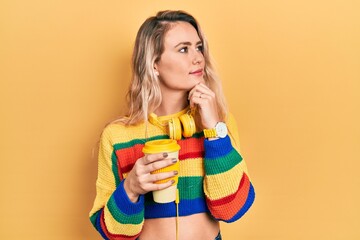 Beautiful young blonde woman drinking cup of coffee wearing headphones with hand on chin thinking about question, pensive expression. smiling with thoughtful face. doubt concept.