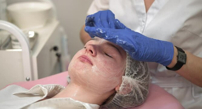 Close up of hands of cosmetologist in blue rubber gloves, applying professional peeling or cleansing foam to young teen girl face. Acne and skin pimple treatment at modern clinic. Problem Skin