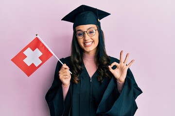 Young hispanic woman wearing graduation uniform holding switzerland flag doing ok sign with fingers, smiling friendly gesturing excellent symbol