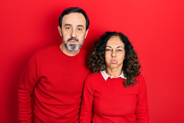 Middle age couple of hispanic woman and man hugging and standing together puffing cheeks with funny face. mouth inflated with air, crazy expression.