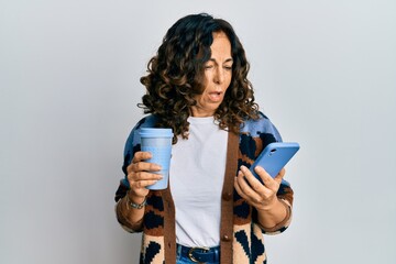 Middle age hispanic woman drinking a cup of coffee and looking at the smartphone screen clueless...