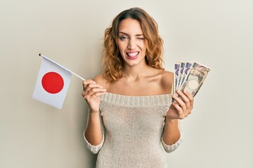 Young caucasian woman holding japan flag and yen banknotes winking looking at the camera with sexy...