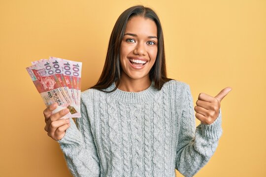 Beautiful hispanic woman holding 100 hong kong dollars banknotes pointing thumb up to the side smiling happy with open mouth