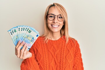 Beautiful blonde woman holding 20 euro banknotes looking positive and happy standing and smiling with a confident smile showing teeth