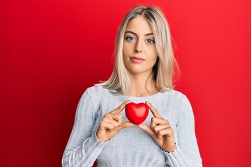 Beautiful blonde woman holding heart relaxed with serious expression on face. simple and natural looking at the camera.
