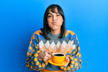 Young hispanic woman drinking a cup of coffee puffing cheeks with funny face. mouth inflated with...