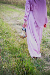 A Muslim woman in a pink hijab walks across a field of wheat with shoes in her hands.