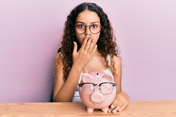 Obraz na płótnie Canvas Teenager hispanic girl holding piggy bank with glasses covering mouth with hand, shocked and afraid for mistake. surprised expression