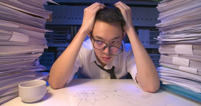 Frustrated asian business man is looking at stack of paperwork, tearing his hair and feeling desperate. Exhausted alone male is sighing and lay down on desk while working hard overload at late night.
