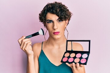 Young man wearing woman make up holding makeup brush and blush skeptic and nervous, frowning upset because of problem. negative person.
