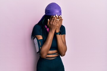 Young african american woman wearing gym clothes and using headphones with sad expression covering face with hands while crying. depression concept.