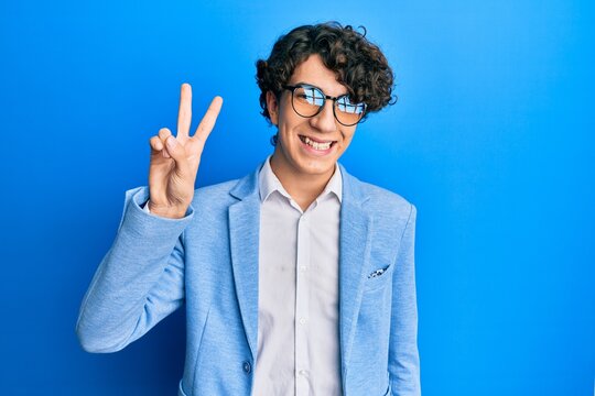 Hispanic young man wearing business jacket and glasses smiling with happy face winking at the camera doing victory sign. number two.