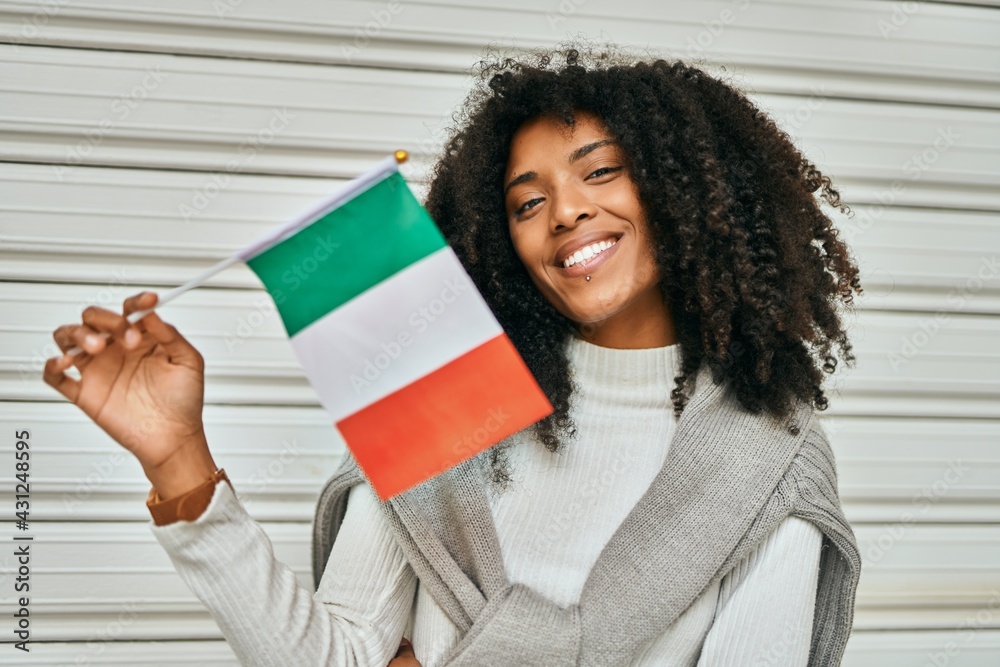 Wall mural Young african american woman smiling happy holding Italy flag at the city.