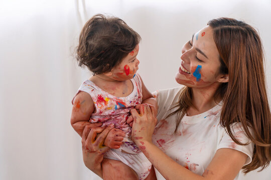 Joyful motherhood hugging own infant daughter with face painting watercolor while playful together relax in the room. Happiness young woman carrying little baby girl sitting play at home.