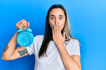 Young hispanic woman holding alarm clock covering mouth with hand, shocked and afraid for mistake. surprised expression