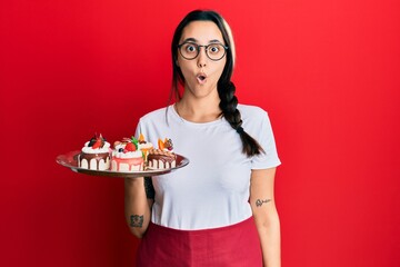 Young hispanic woman wearing waiter uniform holding cakes scared and amazed with open mouth for surprise, disbelief face