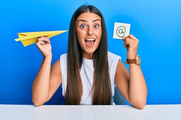 Beautiful hispanic woman holding paper plane and email symbol celebrating crazy and amazed for...