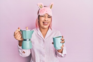 Hispanic woman with pink hair wearing sleep mask and pajama drinking a coffee cup winking looking...