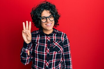 Young hispanic woman with curly hair wearing casual clothes and glasses showing and pointing up with fingers number three while smiling confident and happy.