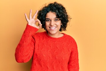 Young hispanic woman with curly hair wearing casual winter sweater smiling positive doing ok sign with hand and fingers. successful expression.