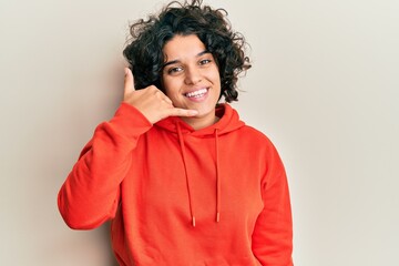Young hispanic woman with curly hair wearing casual sweatshirt smiling doing phone gesture with hand and fingers like talking on the telephone. communicating concepts.