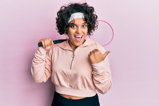 Young hispanic woman with curly hair holding badminton racket pointing thumb up to the side smiling happy with open mouth