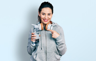 Young hispanic woman wearing sportswear holding water bottle using headphones smiling happy pointing with hand and finger