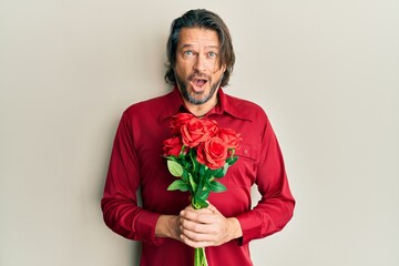 Middle age handsome man holding bouquet of red roses afraid and shocked with surprise and amazed expression, fear and excited face.