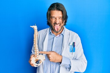 Naklejka premium Middle age handsome traumatologist man holding anatomical model of spinal column sticking tongue out happy with funny expression.