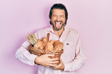 Middle age caucasian man holding wicker basket with bread sticking tongue out happy with funny...