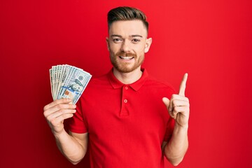 Young redhead man holding dollars smiling with an idea or question pointing finger with happy face, number one