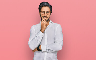 Young hispanic man wearing business shirt and glasses looking confident at the camera with smile with crossed arms and hand raised on chin. thinking positive.