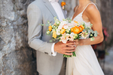 The bride and groom stand hugging at a brick wall in the old city and hold a wedding bouquet with orange flowers, close-up 