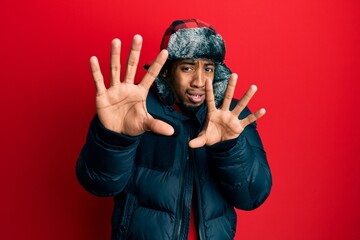 Young african american man with beard wearing winter hat and coat afraid and terrified with fear expression stop gesture with hands, shouting in shock. panic concept.