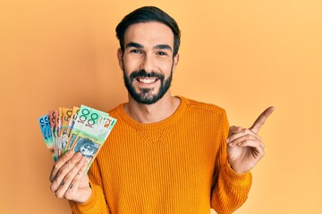 Young hispanic man holding australian dollars smiling happy pointing with hand and finger to the side