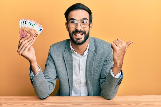 Young hispanic man holding euro banknotes pointing thumb up to the side smiling happy with open mouth