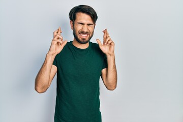 Young hispanic man wearing casual white tshirt gesturing finger crossed smiling with hope and eyes closed. luck and superstitious concept.