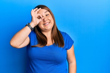 Beautiful brunette plus size woman wearing casual blue t shirt doing ok gesture with hand smiling, eye looking through fingers with happy face.