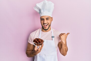 Young arab man wearing professional cook uniform holding bowl with dates pointing thumb up to the side smiling happy with open mouth