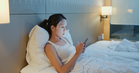 Woman apply face mask and use of mobile phone on bed at night
