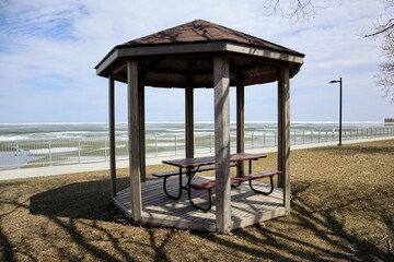 A gazebo with a brown roof, wooden floor, table and benches, standing by the lake.