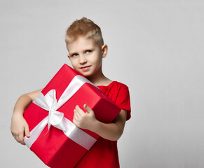 Portrait of cheerful kid boy in red t-shirt holding big present box with ribbon in hands, carrying it over white background with copy space. Trendy casual children fashion concept