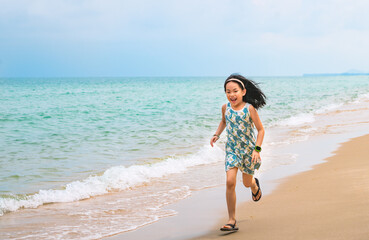 Happy little girl is running along the beautiful beach, motion blur, smiling face, wearing cute dress, black long hair, 7 years old, turquoise color ocean and blue sky. Space for copy and design.