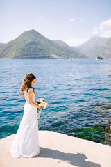 Fototapeta na wymiar An elegant bride with a bouquet in her hands stands on a pier in the Bay of Kotor near the blue transparent water 