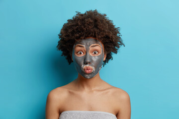 Beauty and skin care concept. Surprised Afro American woman with curly hair applies nourishing clay mask on face enjoys detox treatment wrapped in bath towel shows bare shoulders isolated on blue