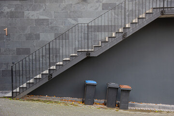 Stairs with steel handrail outside on a wall with gray wall tiles and garbage cans beneath
