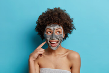 Beauty and cosmetology concept. Happy ethnic curly haired woman smiles pleasantly applies facial clay mask wants to look beautiful wrapped in soft towel isolated over blue studio background.