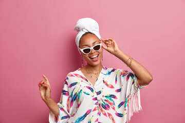 Positive dark skinned woman dressed in domestic gown wears sunglasses bath towel on head feels refreshed after taking shower has healthy skin smiles broadly dances over pink background being at home