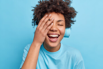 Positive currly haired woman makes face palm smiles happily has carefree expression listens audio track via headphones dressed in casual t shirt isolated over blue background. Emotions lifestyle